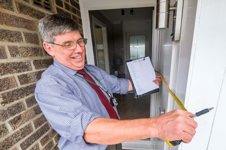 A photograph of a man measuring outside of a doorway with a tape measure